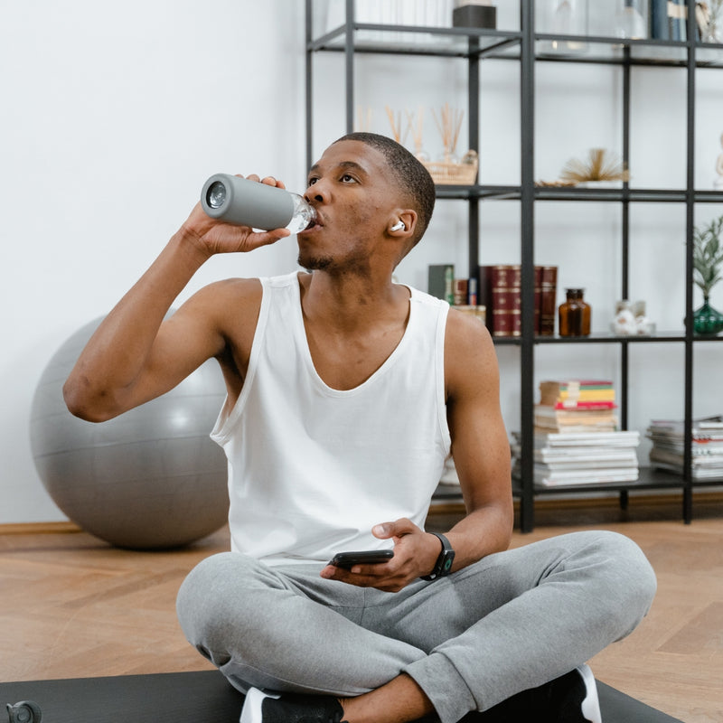 Man sitting on a yoga mat, drinking from a water bottle in a home setting.