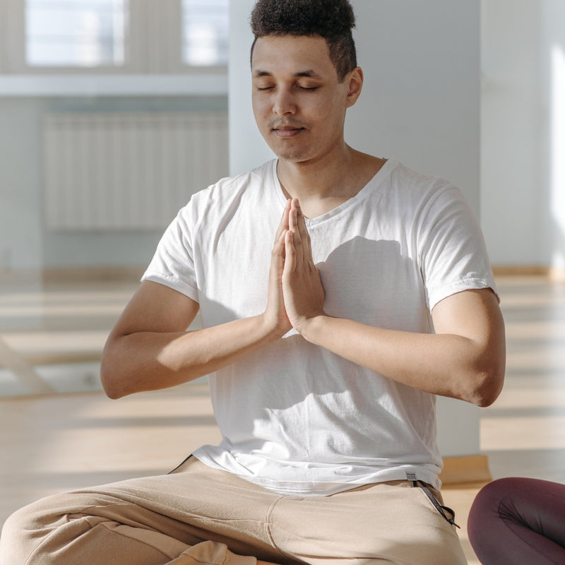 Man in a white t-shirt and beige pants sitting in a meditative pose with hands pressed together.